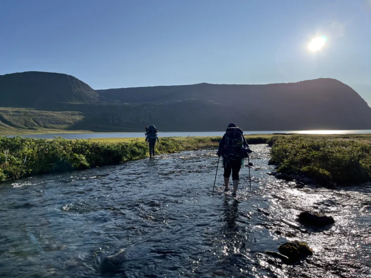 Two people with backpacks walk across a very shallow stream in a field.