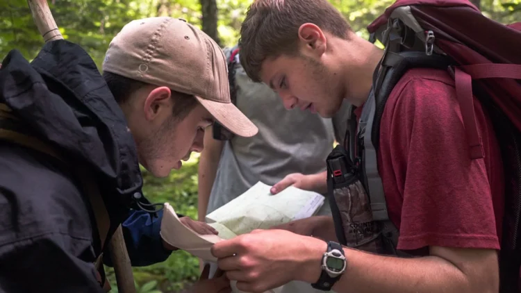 Two young backpackers study a topographic map together in a lush green forest, one wearing a tan baseball cap and dark jacket, the other in a maroon shirt with a hiking pack, practicing navigation skills.