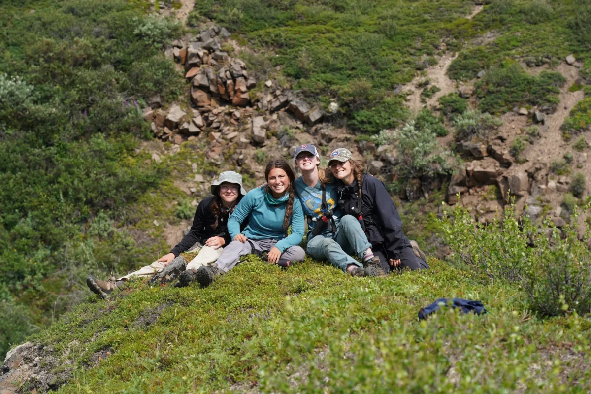 Four hikers sit together on a mossy green ridge, smiling at the camera with rugged rocky terrain and alpine tundra vegetation stretching behind them.