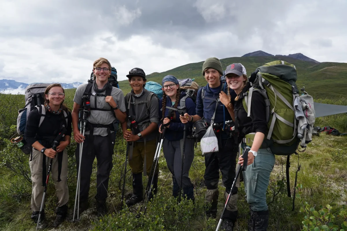 Six smiling backpackers pose together on a hillside in Alaska, wearing full expedition packs and holding trekking poles, with dramatic mountain peaks and moody clouds in the background.