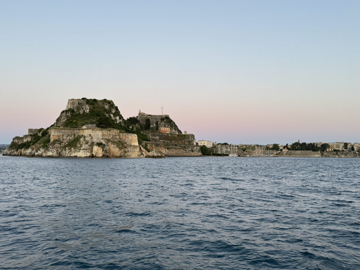 Old fort of Corfu sits on a cliff with ocean in front of it. Skies are a subtle rainbow of colors.