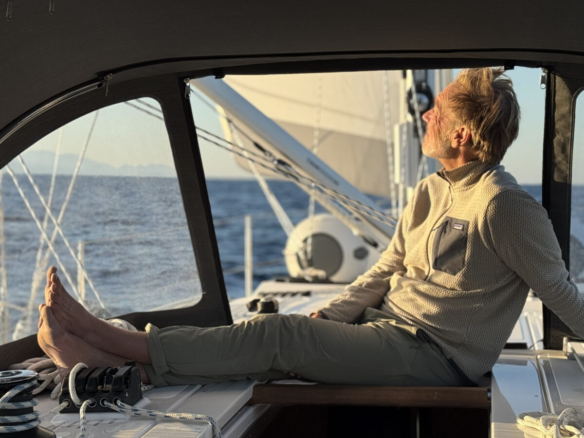 A crew member relaxes on the deck of the sailboat, while the boat is under full sail on the ocean.