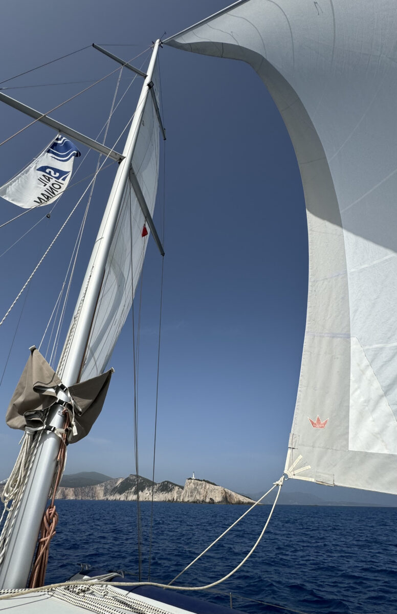 Sails of the boat in full sail on the ocean with cliffs and a lighthouse visible in the background.