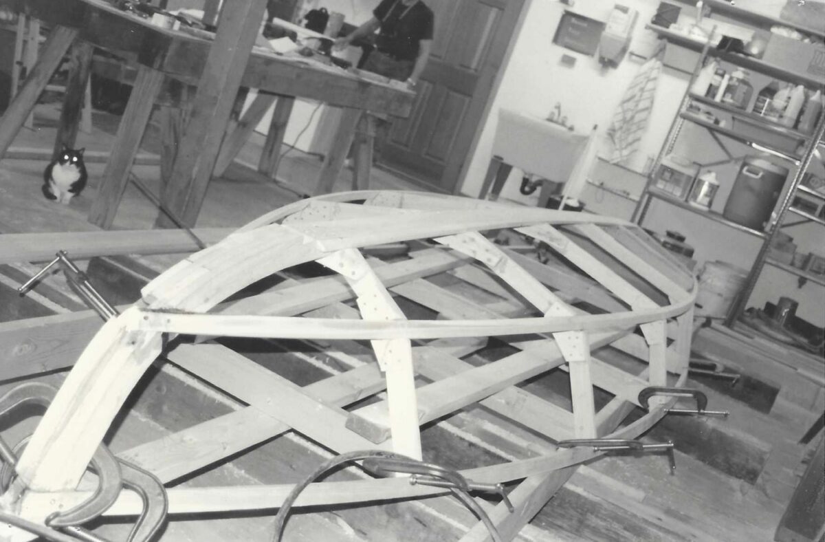 B/W photo of the skeleton of Nick's sailboat in the garage as he and his dad built it. Dad and a cat are partially visible in background.