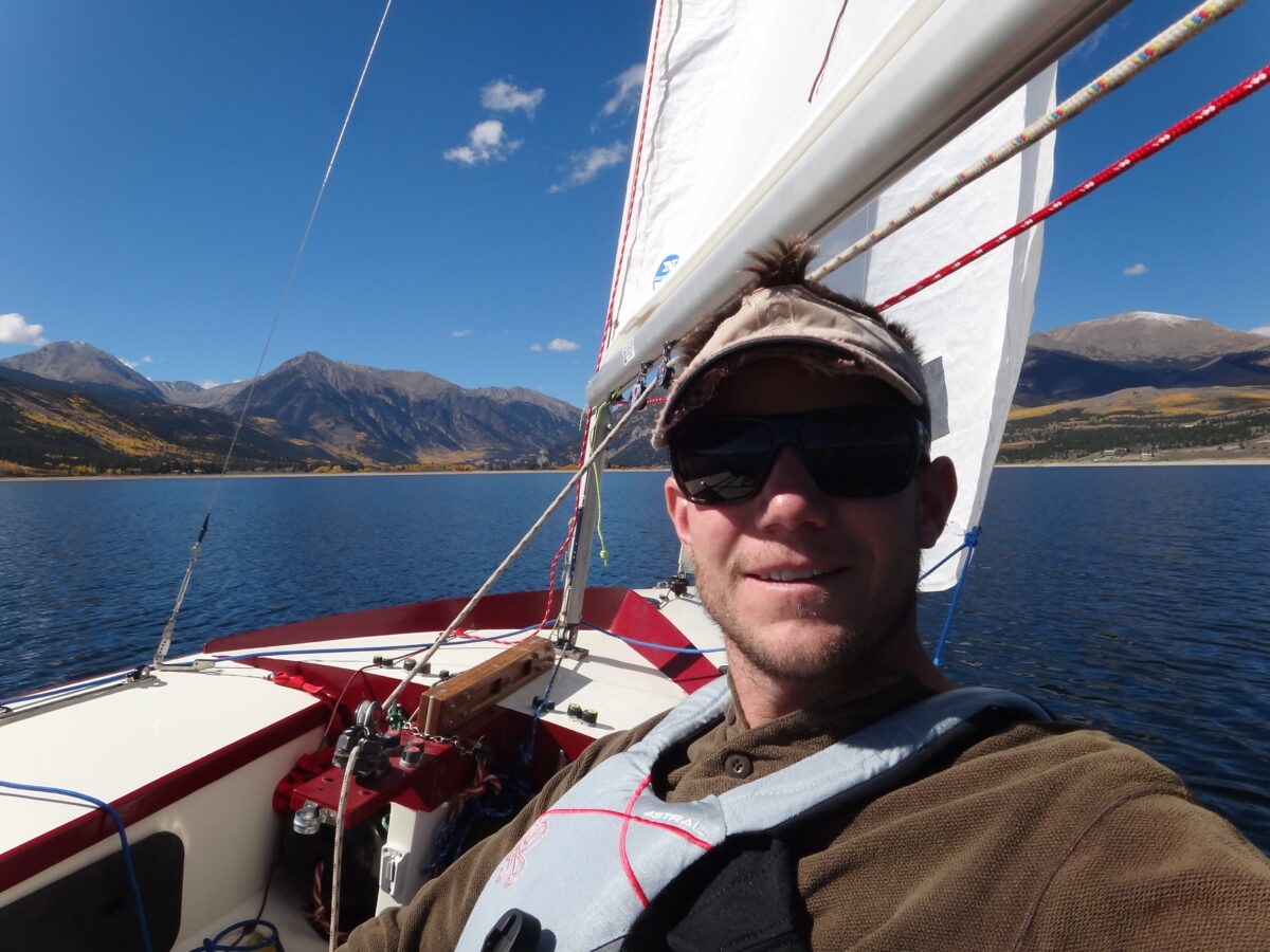 Selfie shot of Nick Braun as he sits aboard the sailboat wearing dark sunglasses and a cap. In the background, there's a blue sky and mountains and a smooth sea.