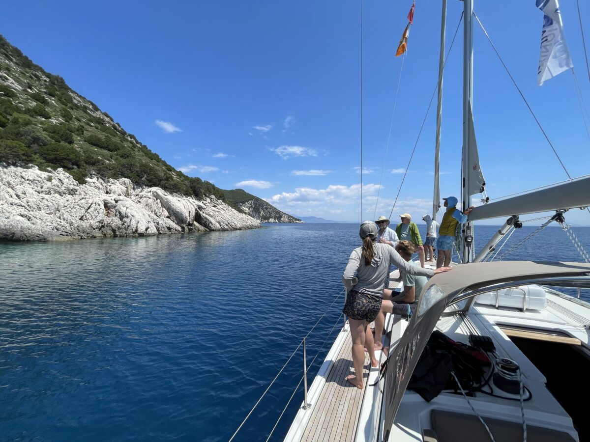 Five person crew stand on the sailboat's deck as they chat. All are watching the cliff to the left.