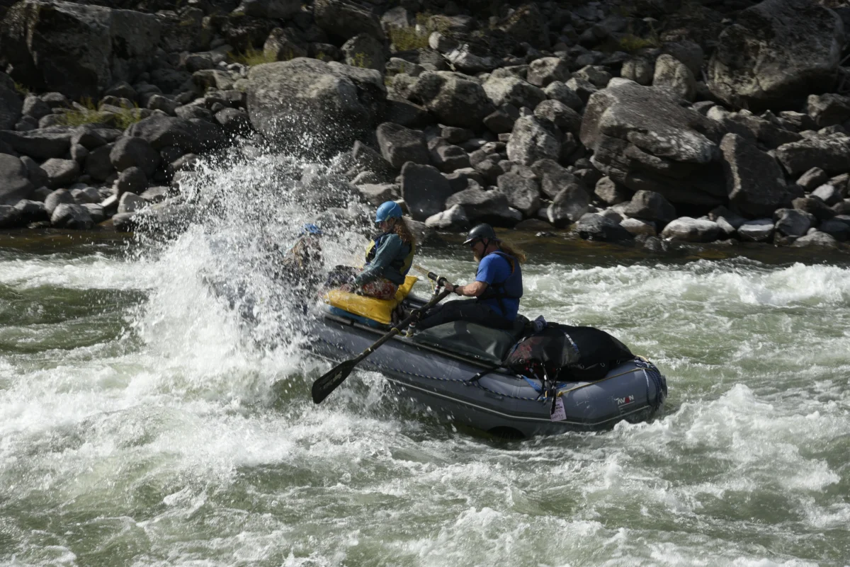 two people in a grey inflatable river raft hit a standing wave. water splashes over the front of the boat.
