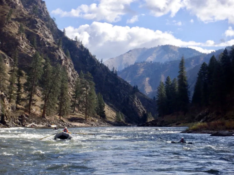 a raft navigates flatwater on a placid river. The hillsides rise steeply from the river. There are mountains in the background.