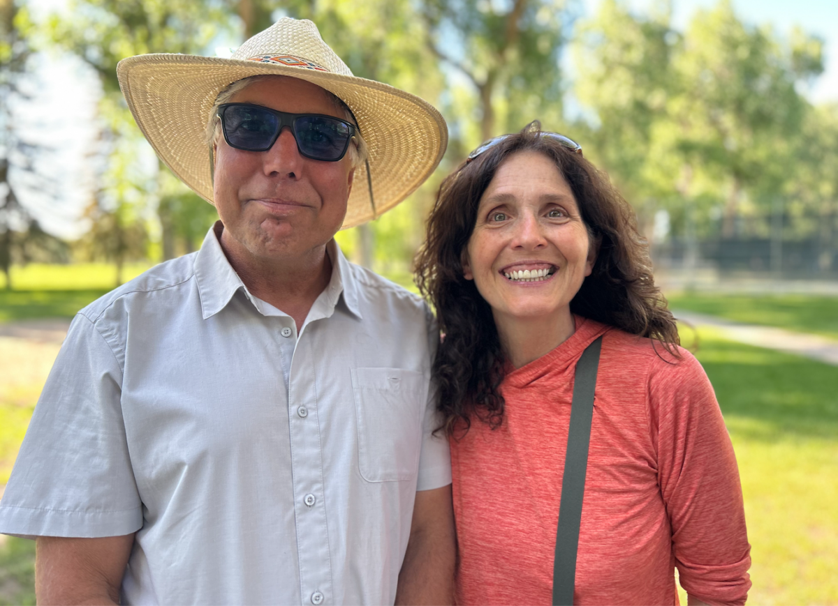 Andy Blair, wearing a sunhat, and Jacki Klancher stand close together  with big smiles on their faces. Green grass and green deciduous trees in the background.