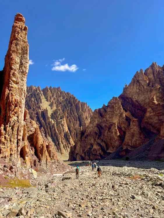 Cliffs rise up from the riverbed with hikers walking away from the camera. Blue skies and sunshine all around.