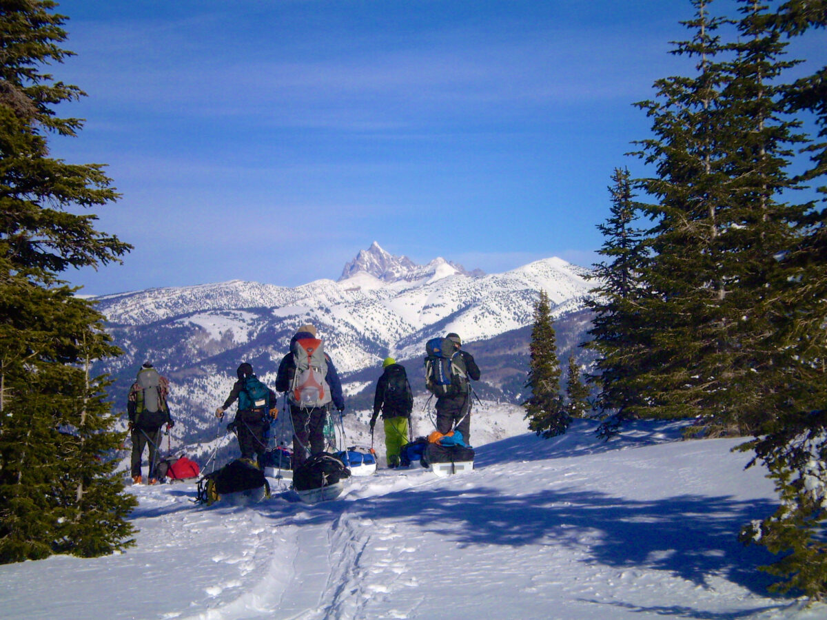 Five skiers wait at the edge of a precipice, beautiful snow-covered mountain peaks and blue skies in the background.
