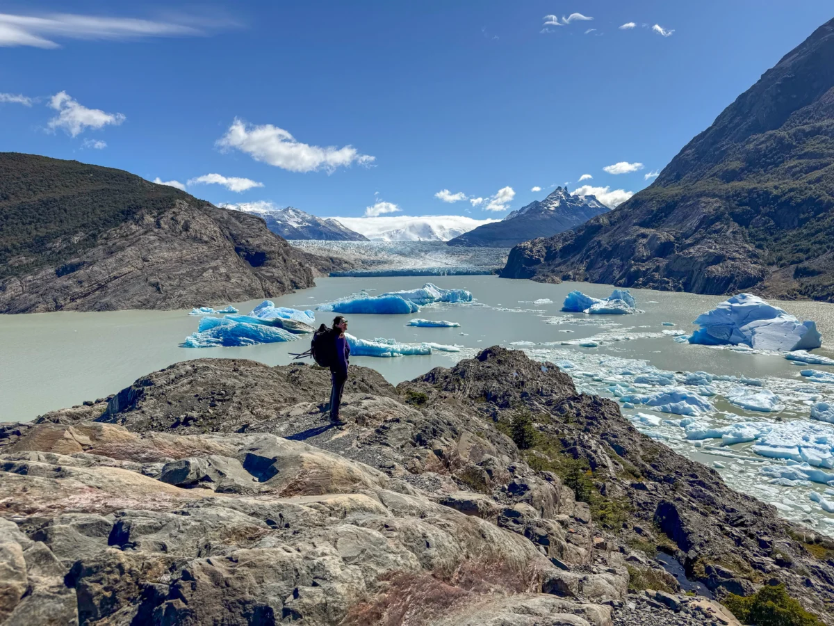 a person stands on a rock outcropping overlooking a turquoise body of water. there are icerbergs floating in the water and mountains flank the image.