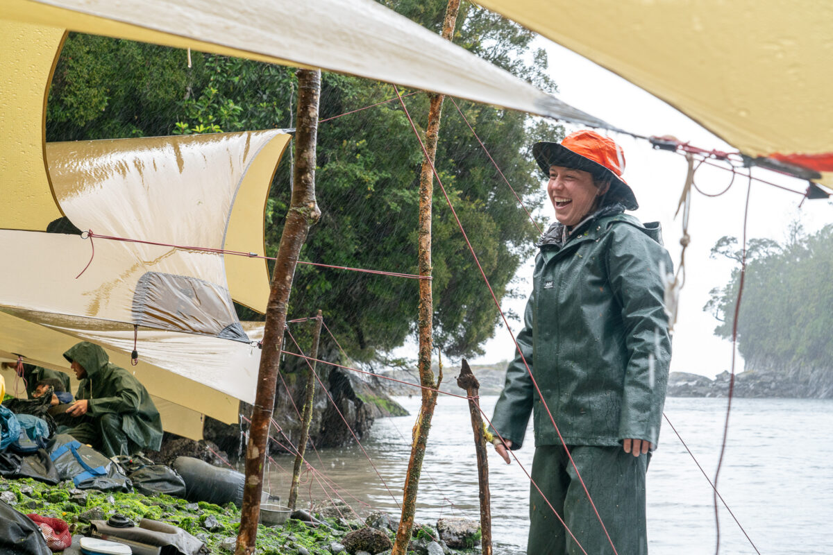 a person wearing green rain gear and an orange rain hat stands in a downpour on the shore. there are yellow tarps set up as shelters but everything is wet