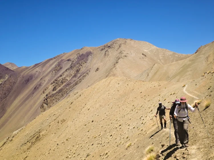 Hikers cross an exposed mountainside with blue skies and sunshine all around.