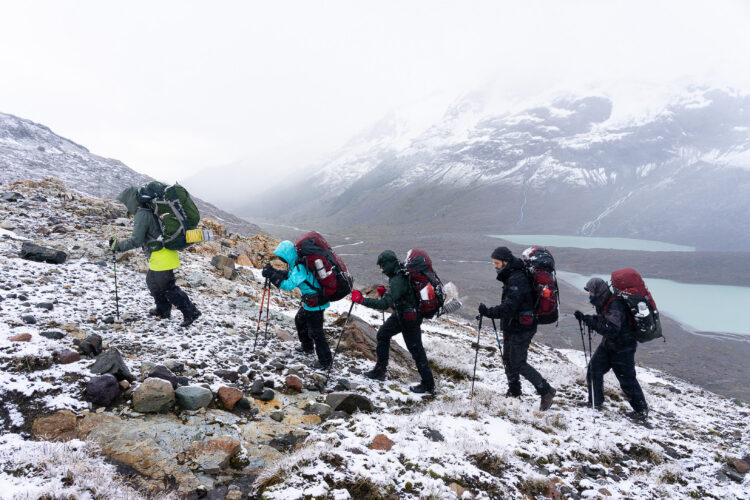 a group of people wearing backpacks hike up a hillside during a snowstorm