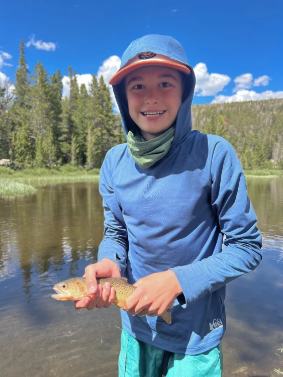 Boy holds fish with lake behind him.