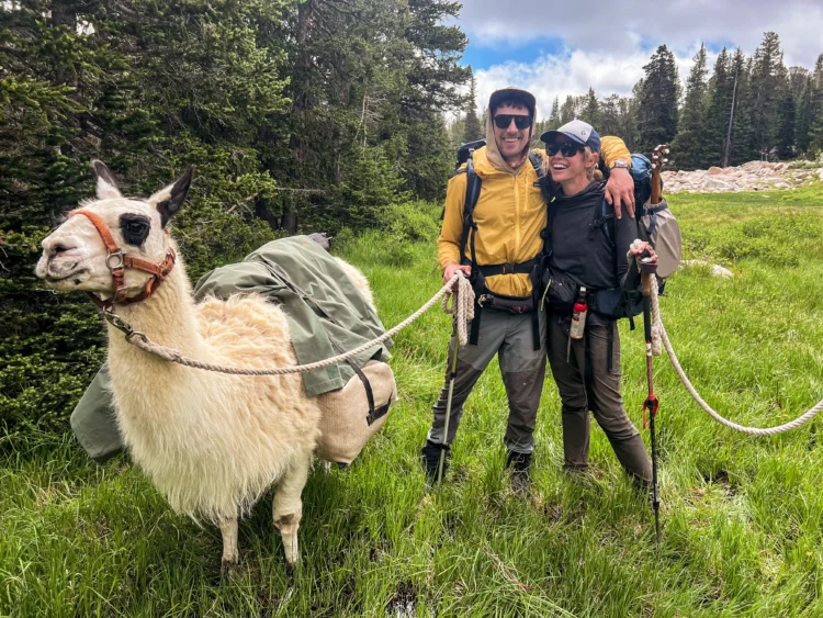Two hikers smile while holding their llama with a rope.