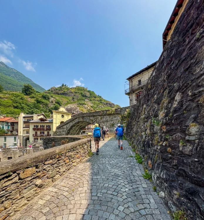 Hikers walk on cobblestone path towards a bridge.