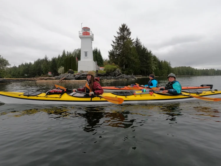 three people in sea kayaks smile at the camera while paddling by a white lighthouse.