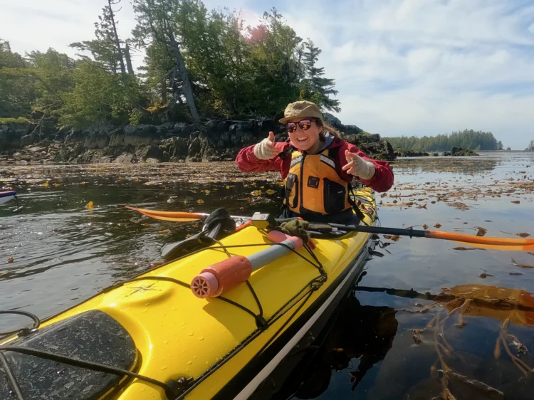 a woman in a yellow sea kayak poses mid-paddle with thumbs up for the camera.
