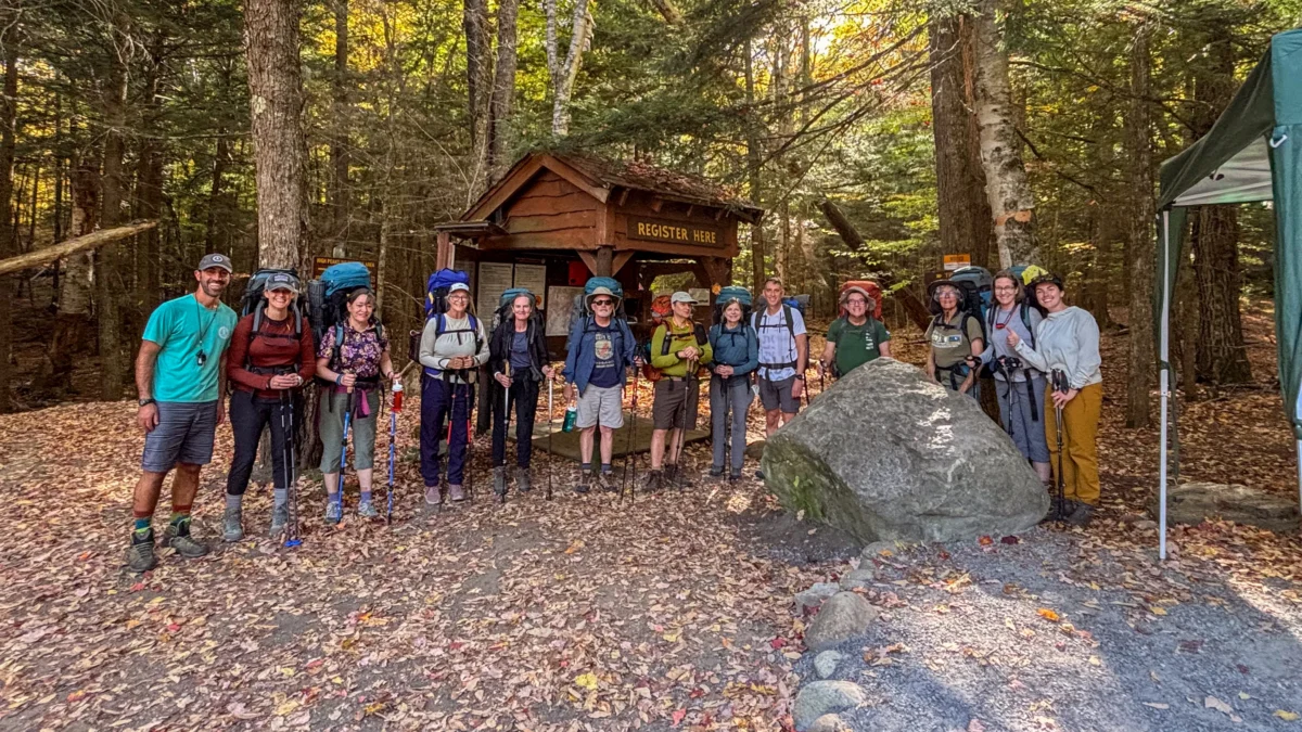 A group of adults pose in a forest by a trail head sign. They are all wearing backpacks.