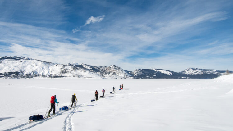 A line of people ski across an open expanse of snow. there are tall mountains in the background. the sky is blue.