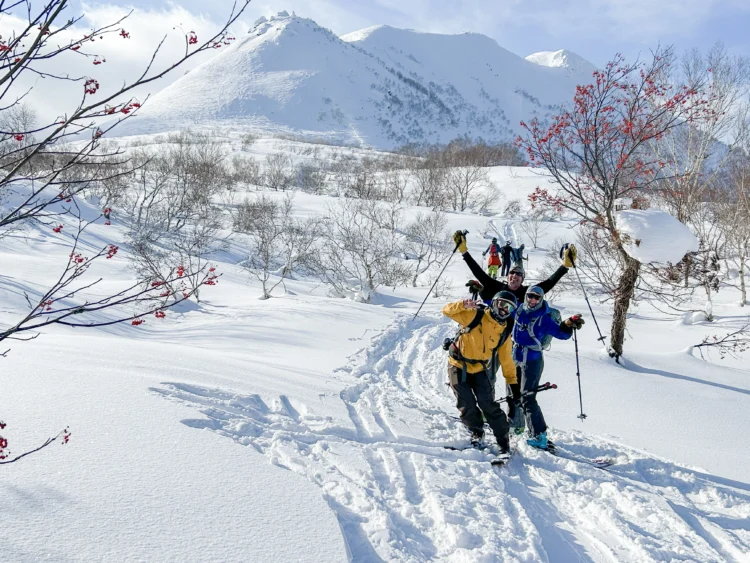 Skiers raise their hands and smile with snow capped mountains behind them.