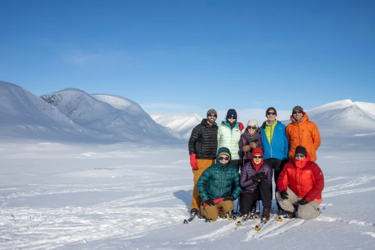 Skiers wearing colorful jackets smile with blue skies and snowcapped mountains behind them. 