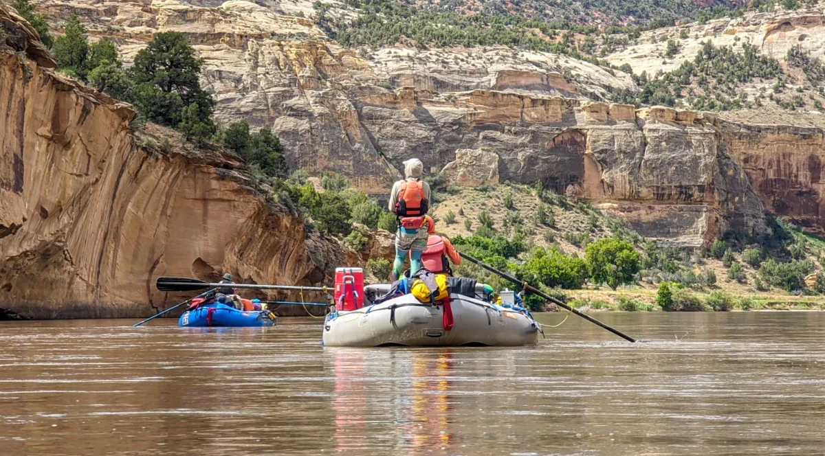 a person wearing a PFD and tights stands in a grey inflatable raft on a still river. There are canyon walls on all sides