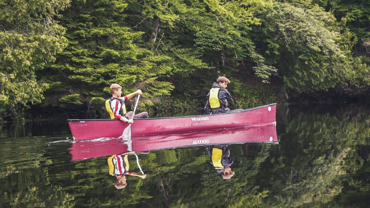 Two teens paddling a red canoe on a calm, tree-lined lake.