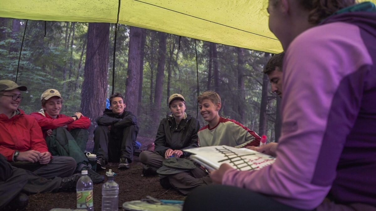 Group of teens sitting together under a tent tarp in the forest during a group therapy session outdoors.
