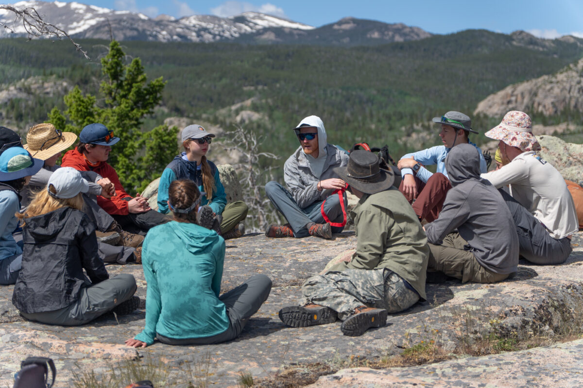 a group of 8 people sit in a circle, talking to one another, on a granite rock. there are mountains in the background. 