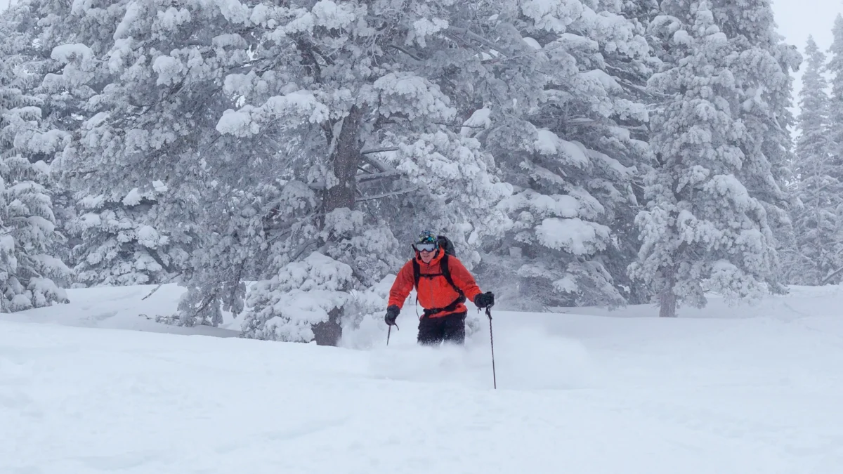 A person in a red coat skis through deep powder against a snowy background.