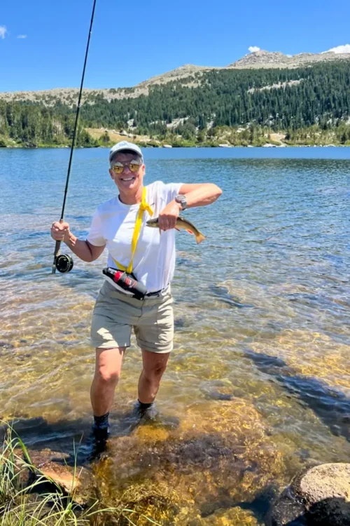 a man in a hat and sunglasses holds up a fish in one hand and a fishing rod in the other. He's standing in a blue lake and there are mountains in the background
