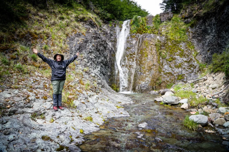 a person in rain gear poses with a smile beside a small waterfall.