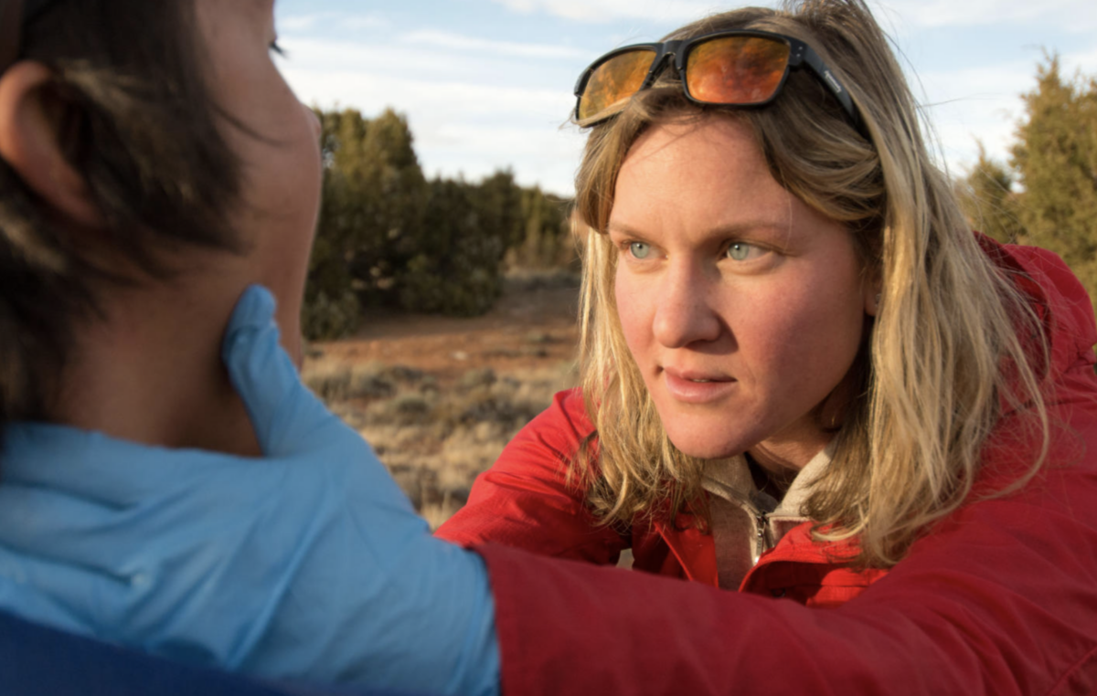 Woman in red jacket and wearing surgical gloves examines a person's face.
