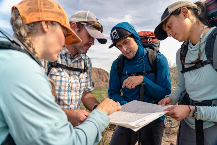 four people wearing baseball hats, sunshirts, and backpacks look at a map
