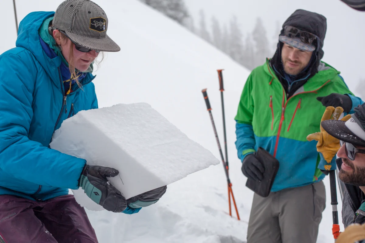 a woman in a blue winter jacket holds a slab of snow in her gloves. two people are looking at the snow slab