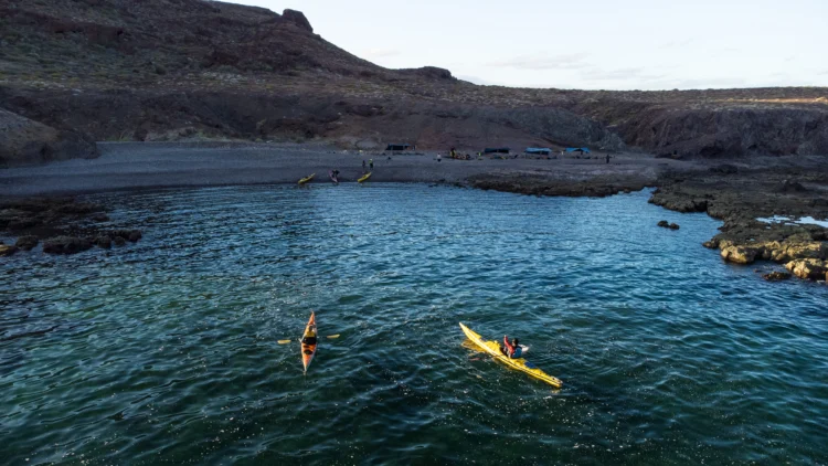 a red and a yellow sea kayak float in a turquoise sea on the edge of a coastline. there are tarps and people on the beach