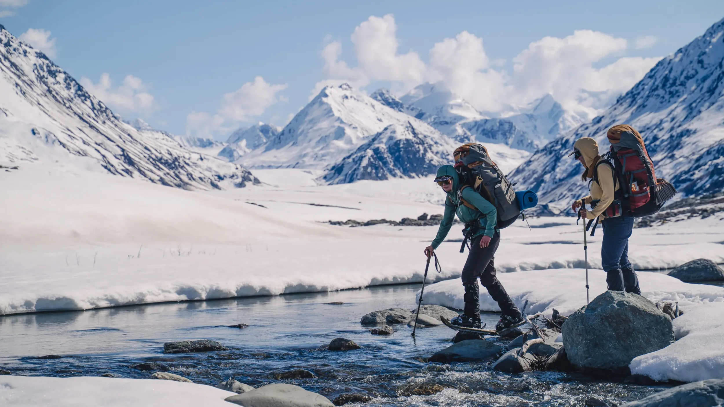 two people wearing snow shoes and large backpacks begin crossing a small stream. there is snow all around