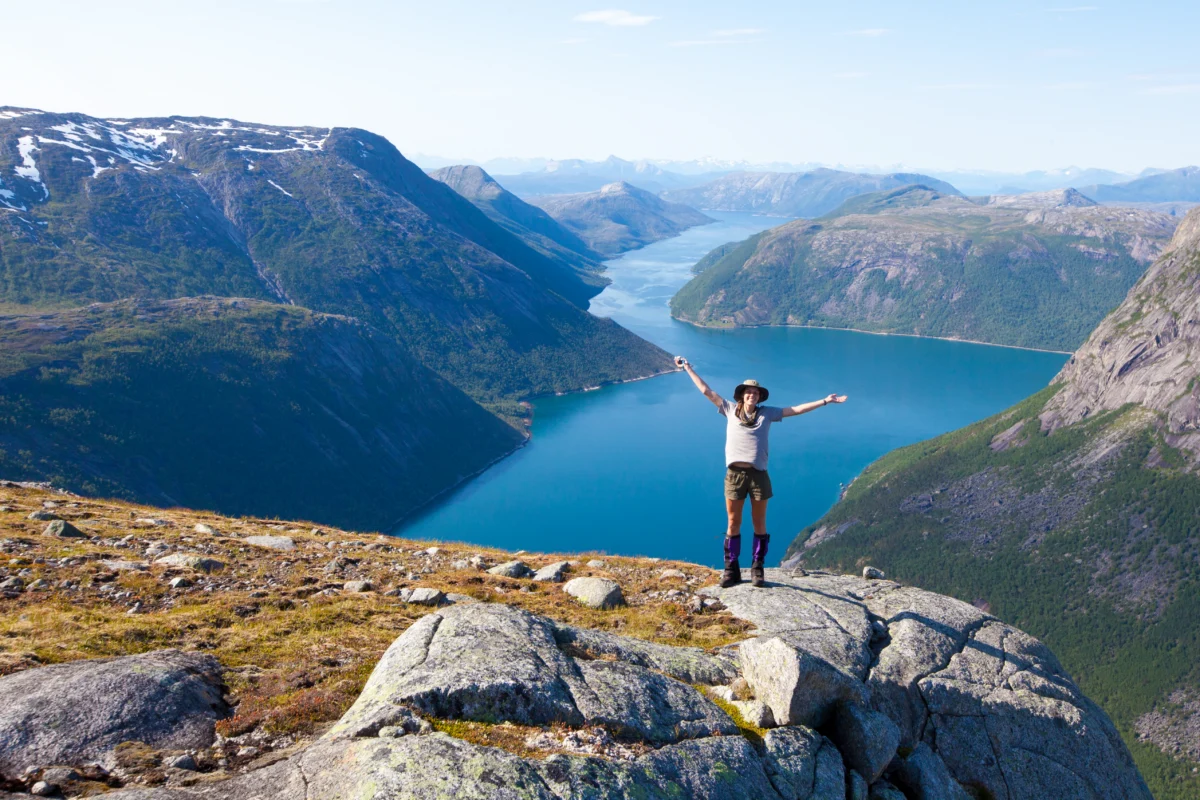 a woman in shorts and pants poses with her arms up in front of a large bright blue fjord