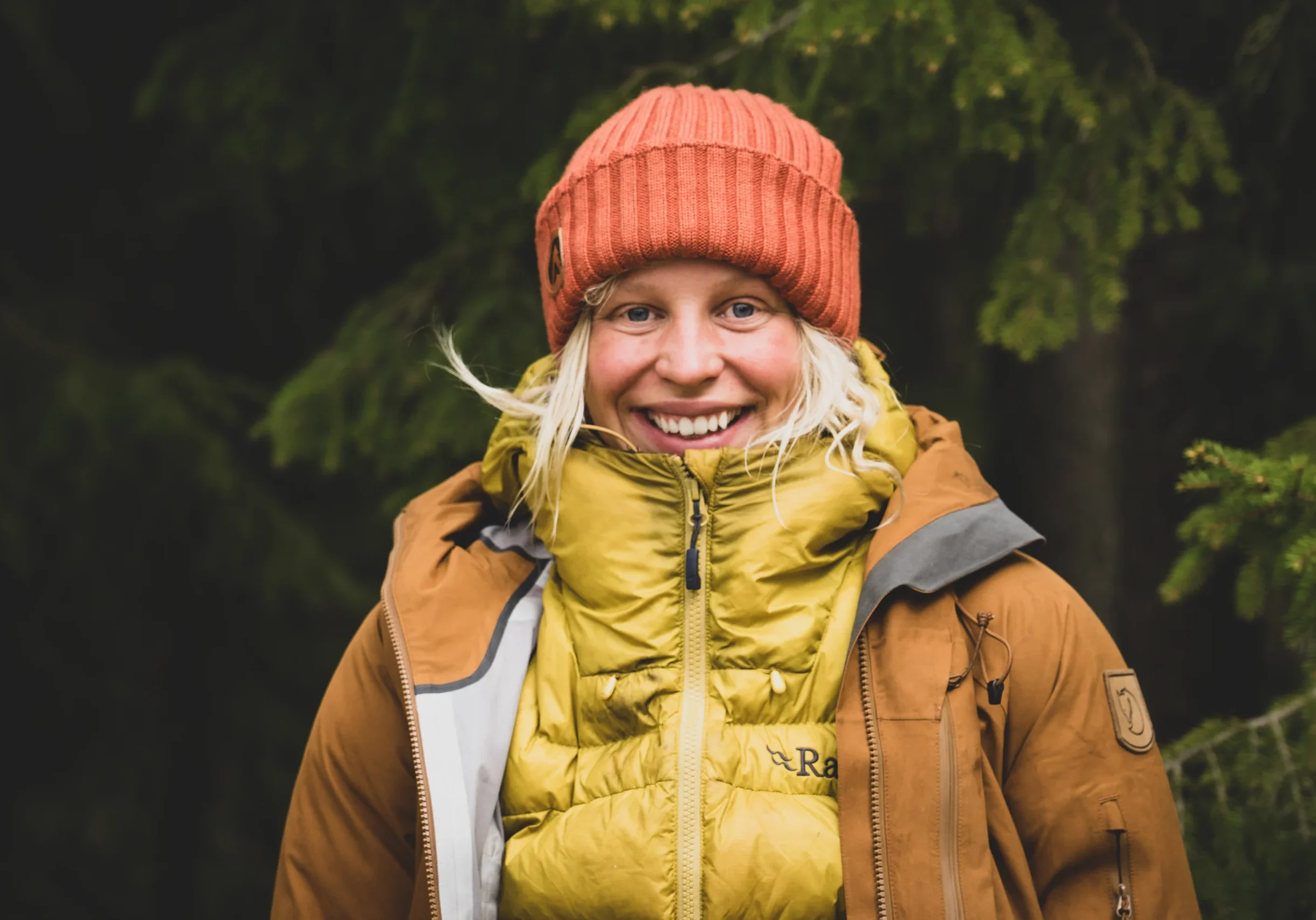 a woman with blond hair wearing an orange winter beanie ad a bright yellow puffy vest smiles widely at the camera