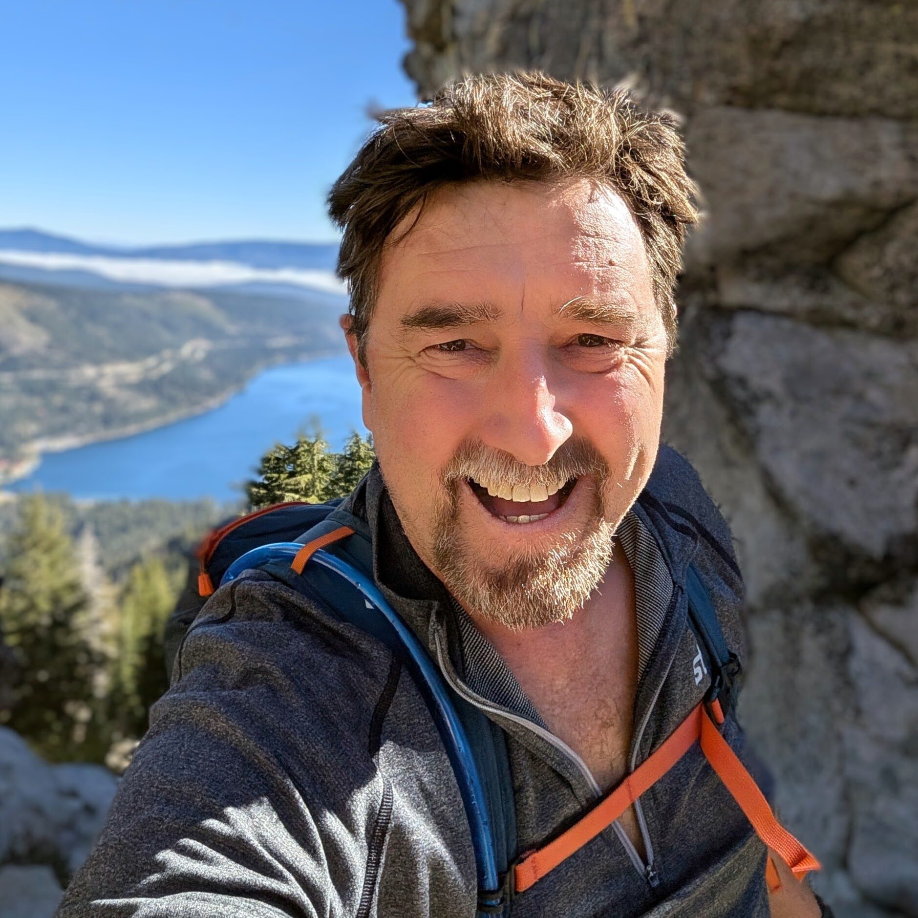 a man smiles at the camera. there is a lake and mountains way below him in the background