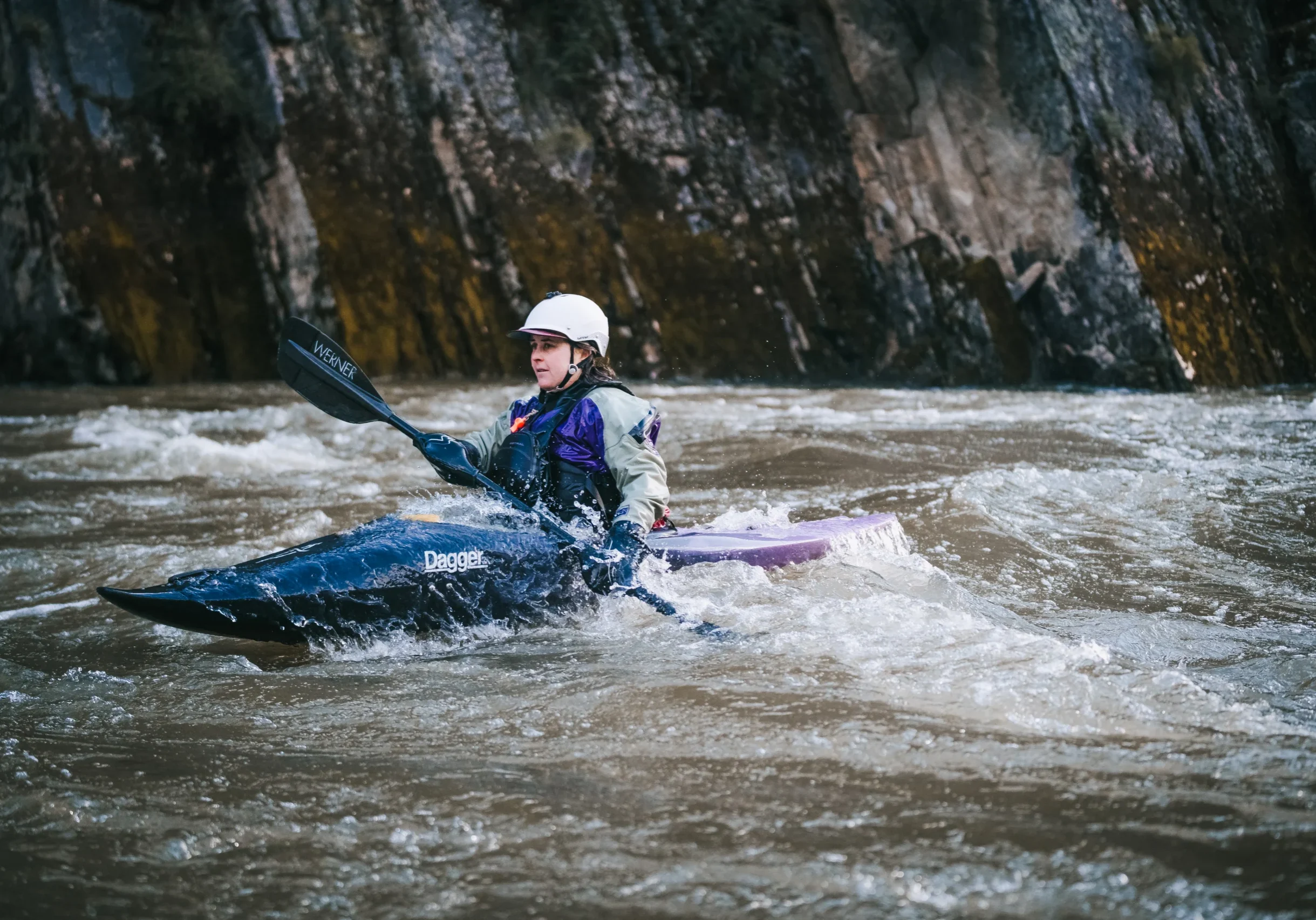 a woman wearing a white helmets paddles a river kayak down a grey river