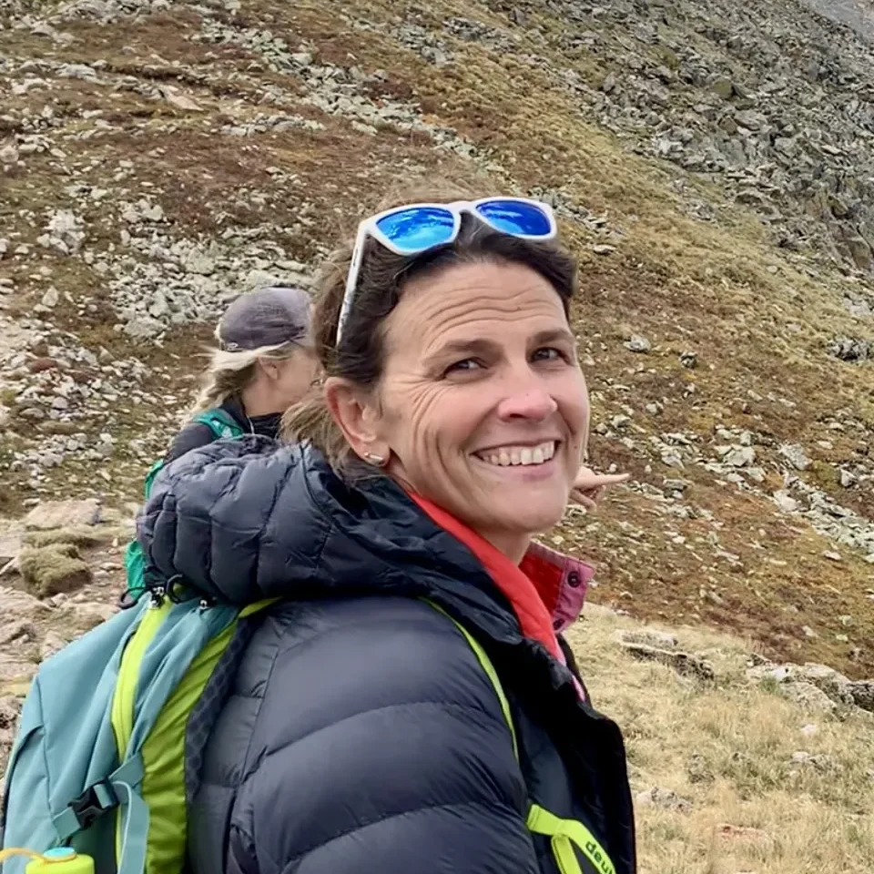 A woman with brown hair and blue sunglasses stands against a grassy mountainside. She is wearing a blue puffy jacket and is smiling at the camera.
