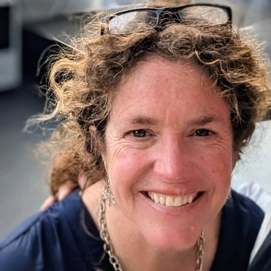 a woman with curly hairs in a ponytail and glasses on her head smiles at the camera.