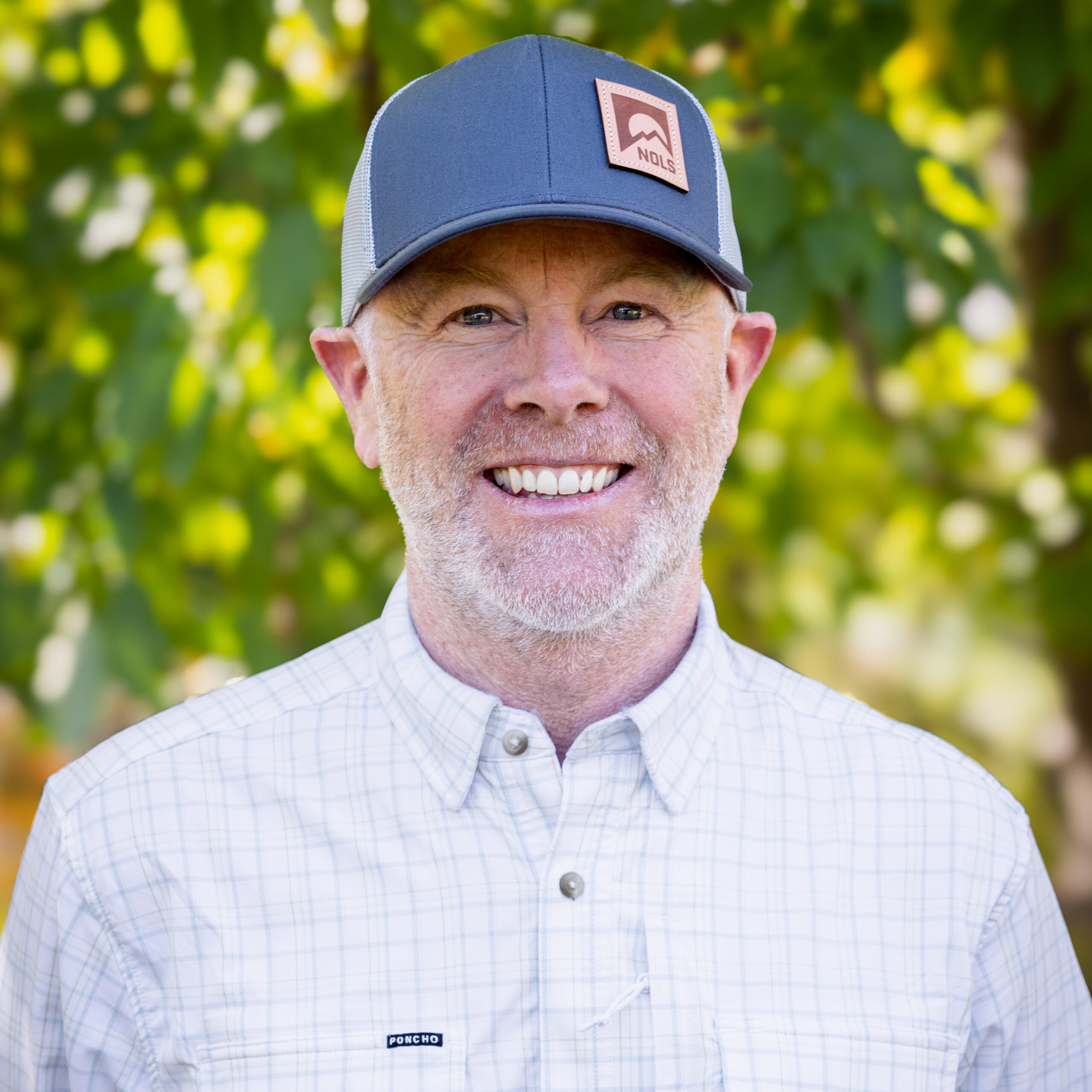 Sandy_Colhoun_headshot_2025 A man wearing a white button down shirt and a trucker's hat that has a NOLS logo smiles for the camera